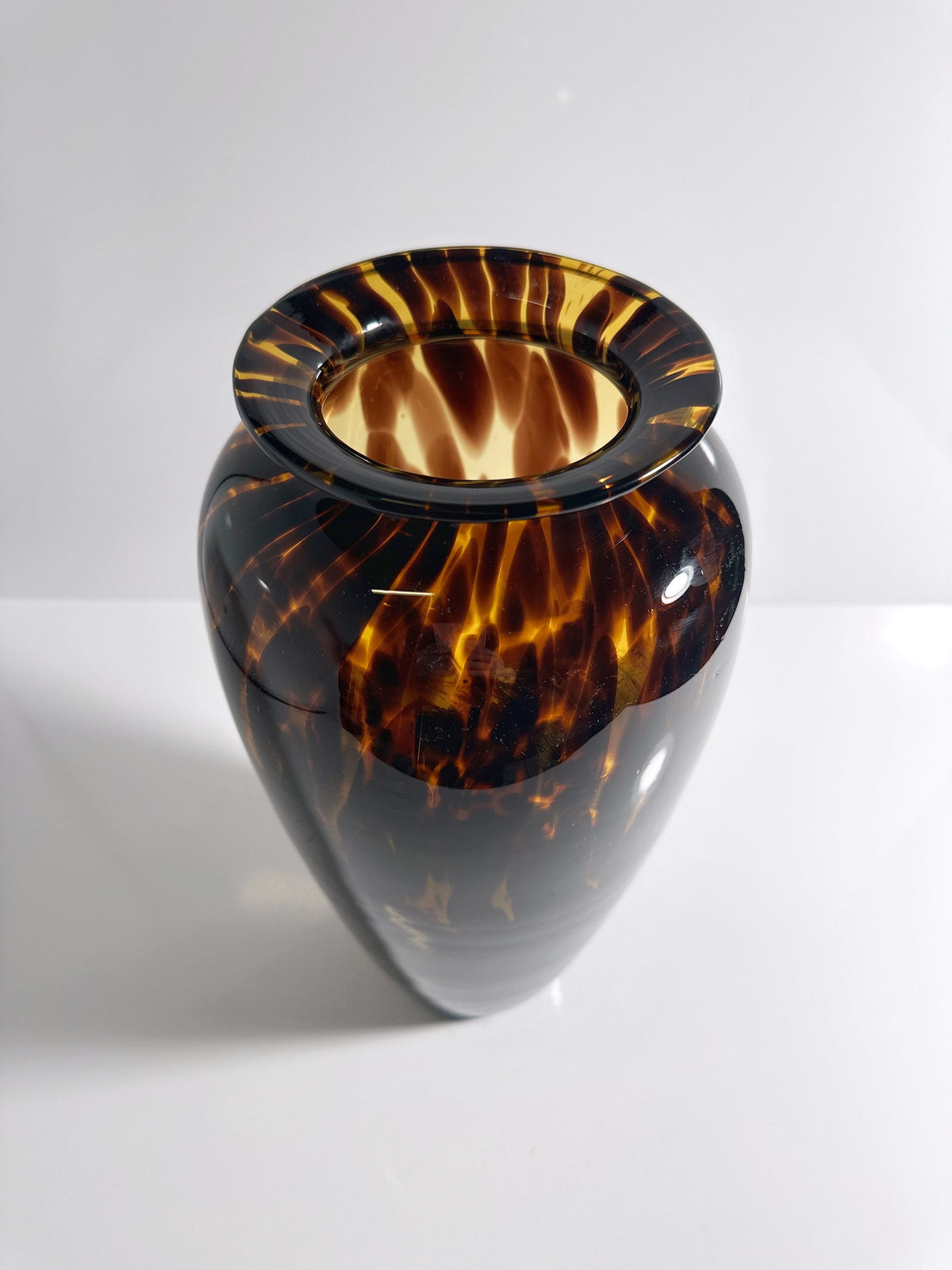 The Tall Murano Tortoise Vase by Dodo Vintage, with its glossy dark brown, tortoiseshell pattern and wide lip, graces the white surface. Its rounded shape and intricate swirling patterns showcase expert craftsmanship in a smooth finish that reflects light.