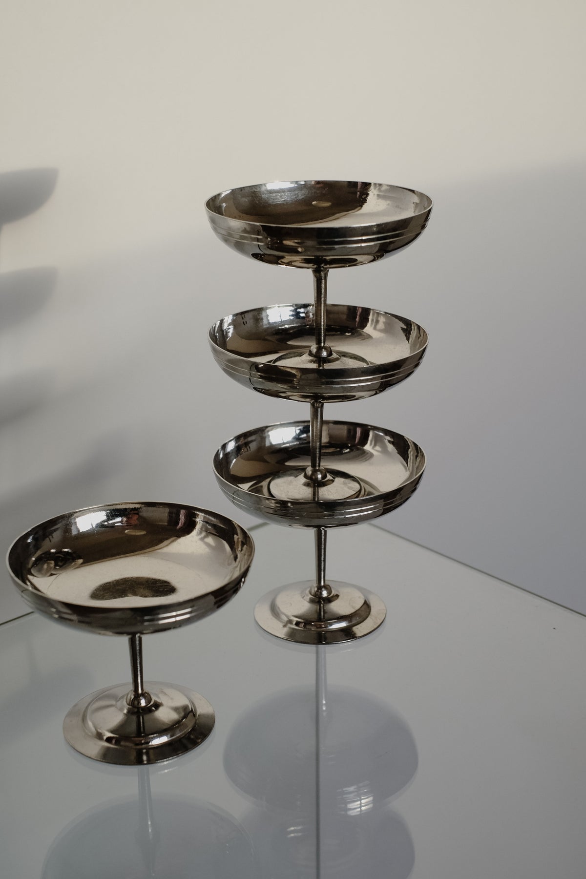A set of four Ice Coupes by septembre studios, crafted from stainless steel and made in France, is stacked on glass with one bowl separately casting shadows on a light-colored background.