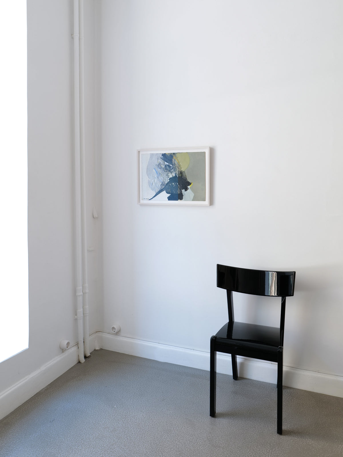 A minimalist room showcases a black chair on a gray carpet against a white wall. Above the chair hangs the "Abstract Composition" painting by Collection apart, featuring shades of blue, gray, and yellow. The space is illuminated by ample light streaming through a large window on the left.