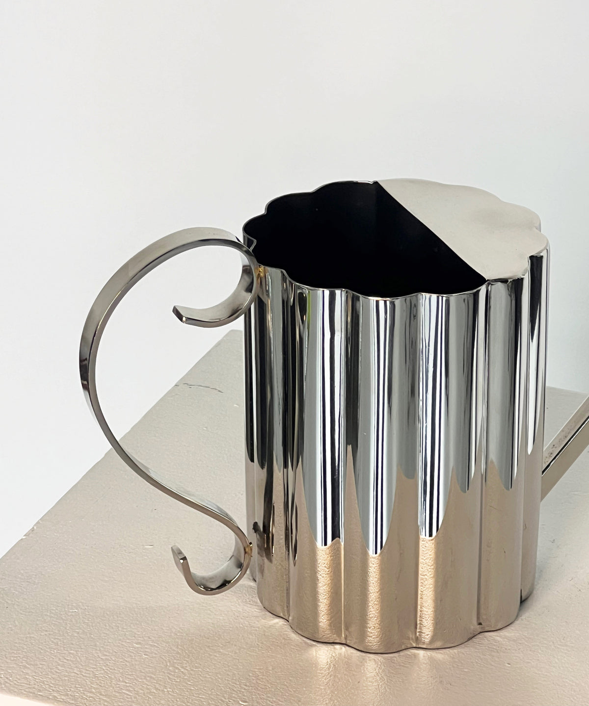 The Fluted Watering Can by Porter Studio is a modern, reflective, silver-plated watering can with a ribbed design and elegant curved handle. It has a triangular spout and sits on a light-colored surface.