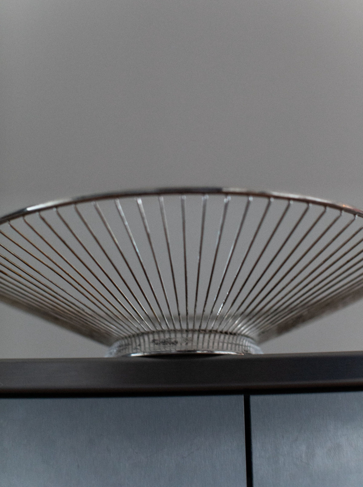 An "Out For Lunch" String Bowl, with its elegant conical shape and evenly spaced thin steel strings, is placed on a gray cabinet against a plain background, creating a symmetric design.