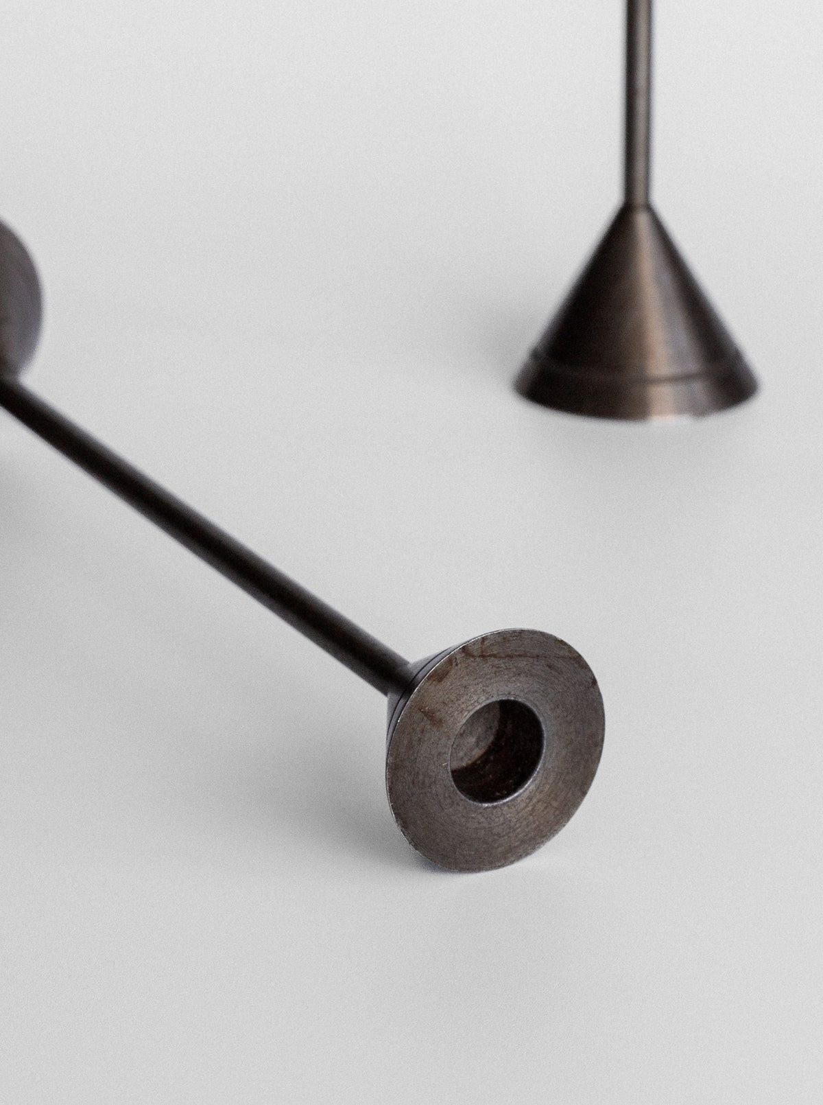 A close-up view of two candlesticks from the Spigel Set of Four Postmodern Candle Holders, lying on a white surface. The sleek metal designs feature cylindrical rods with circular flat bases, one positioned horizontally and the other standing upright. Each base includes a circular cutout in the center, highlighting their unique postmodern style.