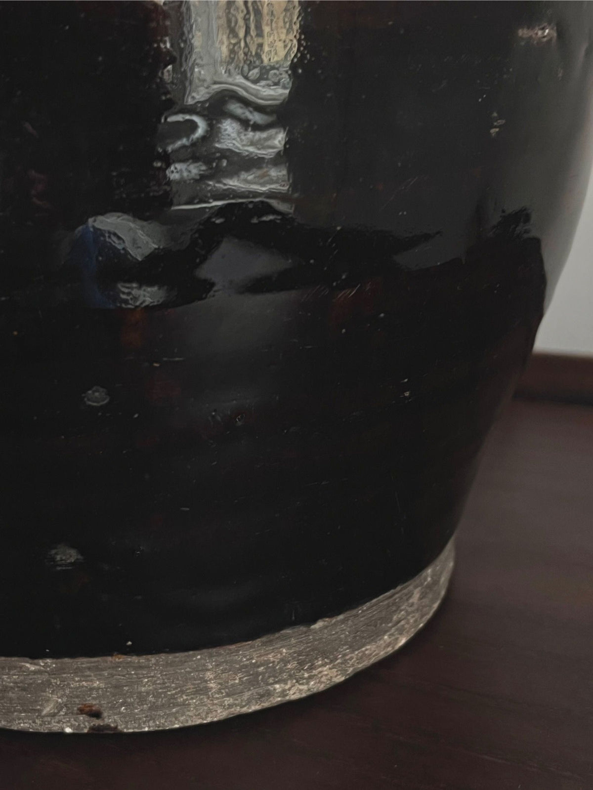 A close-up of the Antique Glazed Lamp by Médecine shows its dark, glossy ceramic body with a metallic silver base. Light dances across its textured, glazed surface, set against a wooden background that contrasts elegantly with the black textile cord draped beside it.
