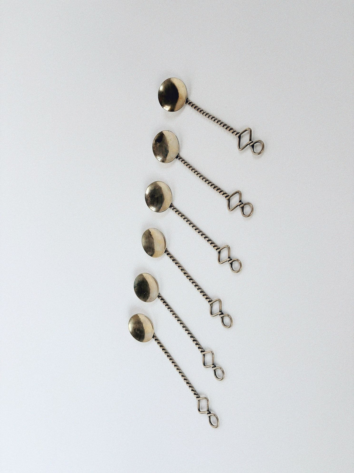 The Huerto 70's Handmade Spoons Set includes six spoons, each with a twisted handle ending in a square loop, arranged vertically on a white background. These functional art pieces seamlessly blend form and practicality, reflecting hints of communist-era design.
