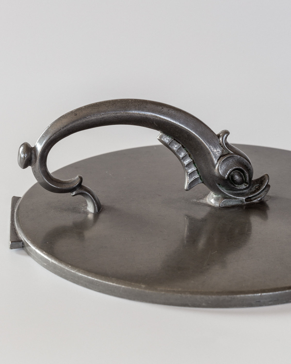 The Art Deco Metal Mirror, created by Spigel, boasts a metal lid with a handle uniquely crafted in the shape of a curved fish or sea creature. Its polished surface artfully reflects light, showcasing elegant craftsmanship that echoes the style of Art Deco mirror designs.