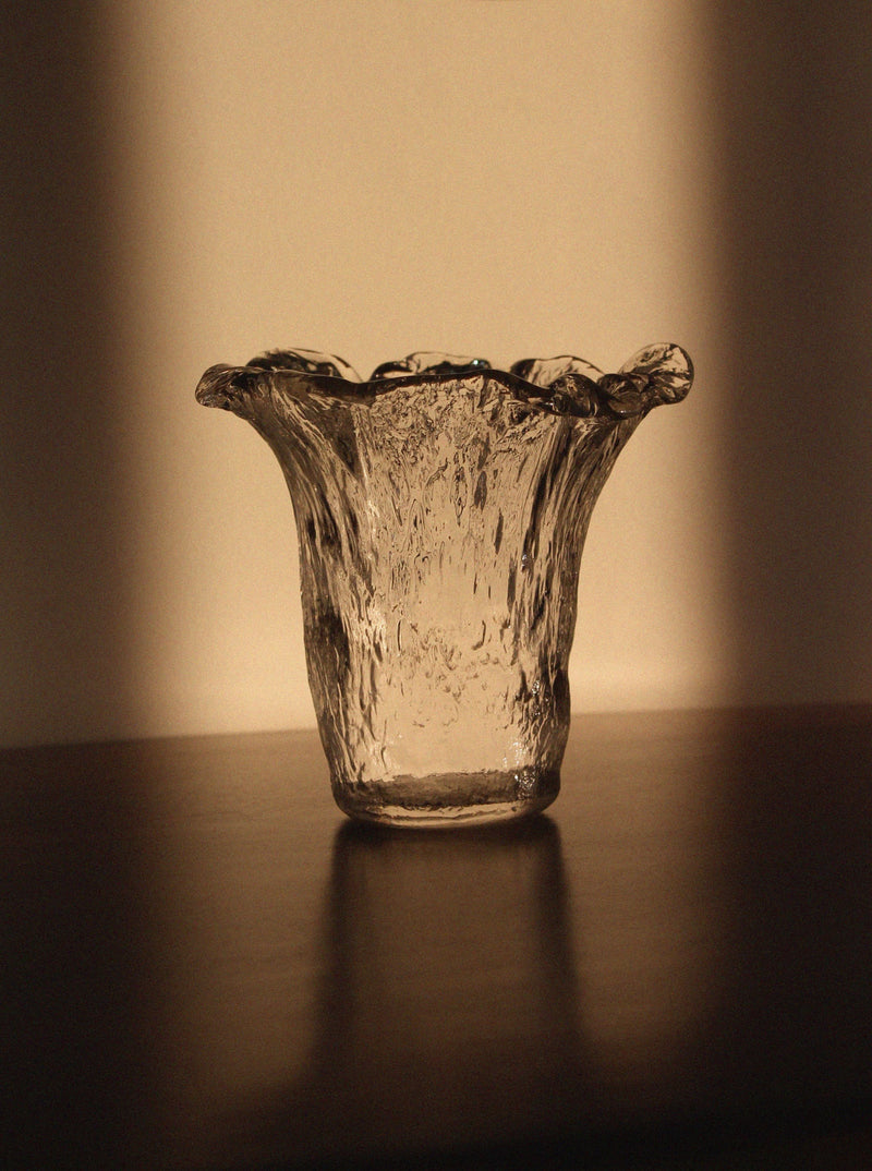 A Uno Westerberg 60s Organic Glass Vase by Vintage Stories, featuring clear textured glass and a flared wavy rim, sits on a dark surface illuminated by warm light that casts gentle shadows in the background.