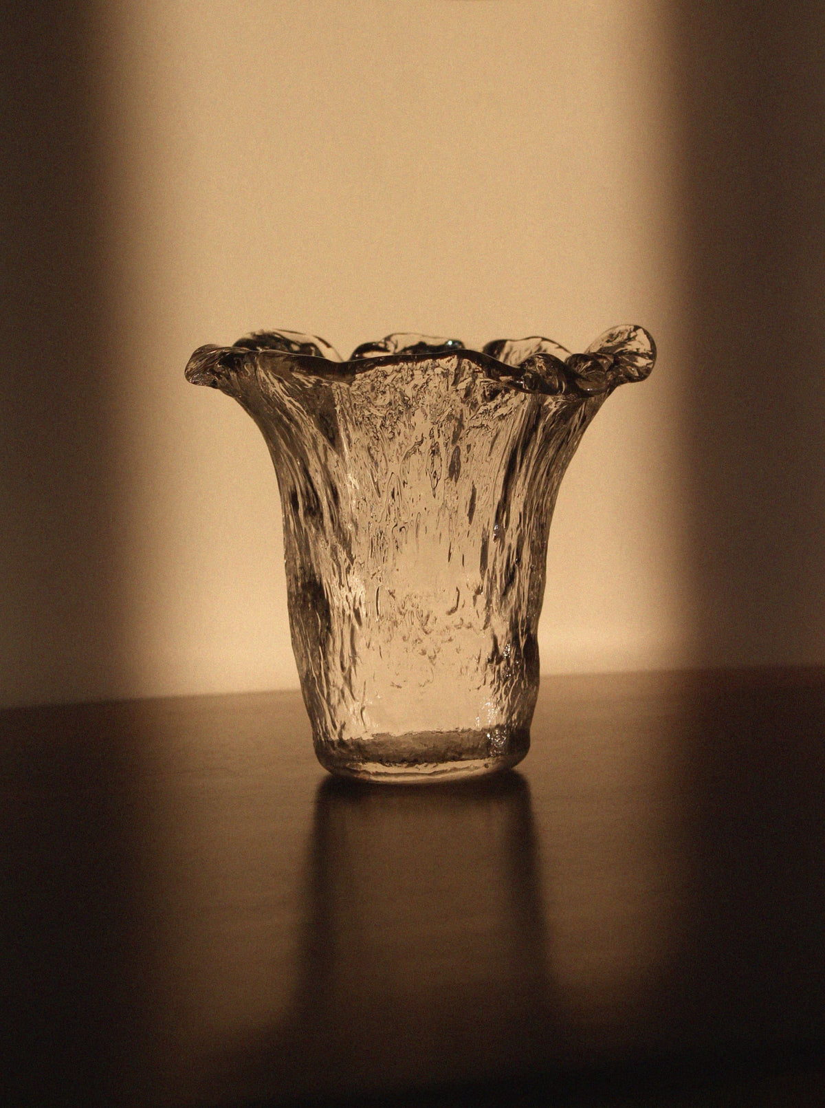 A Uno Westerberg 60s Organic Glass Vase by Vintage Stories, featuring clear textured glass and a flared wavy rim, sits on a dark surface illuminated by warm light that casts gentle shadows in the background.