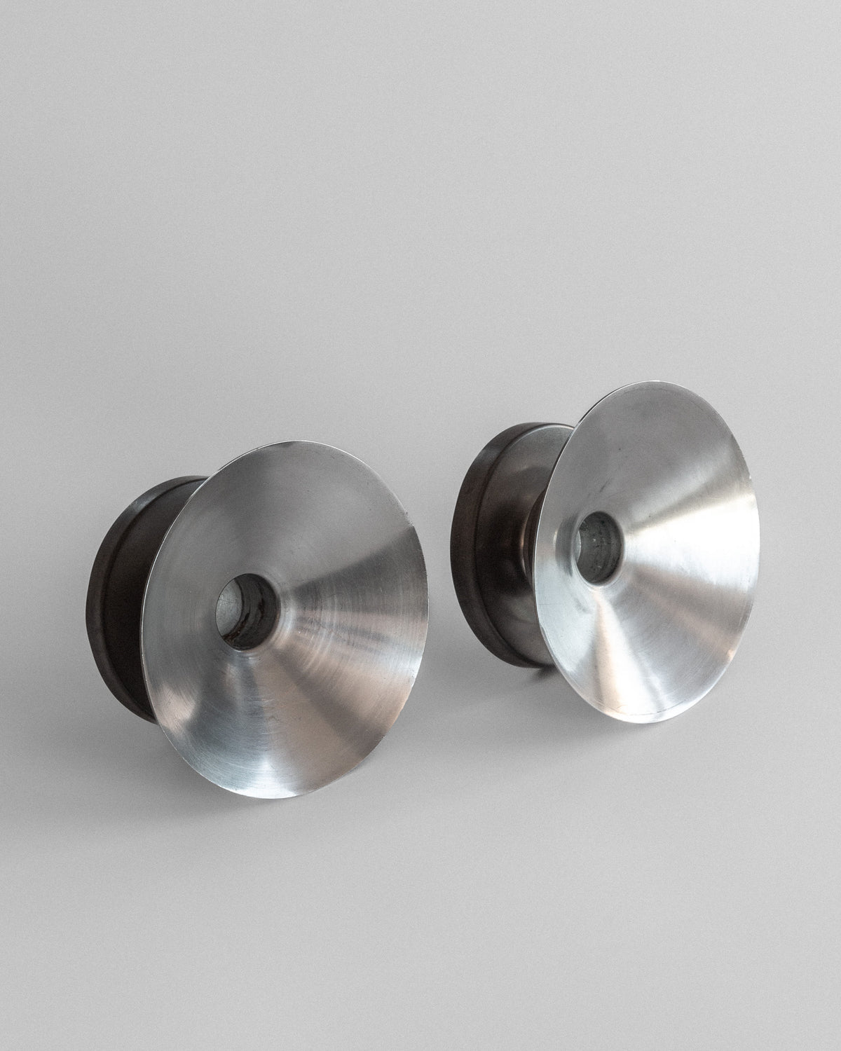 A pair of shiny, silver, conical Modernist Candleholders by Sylvia Stave, created in the 1930s for Spigel, evokes 20th-century metalworks with central holes and reflective symmetry against a plain gray background.