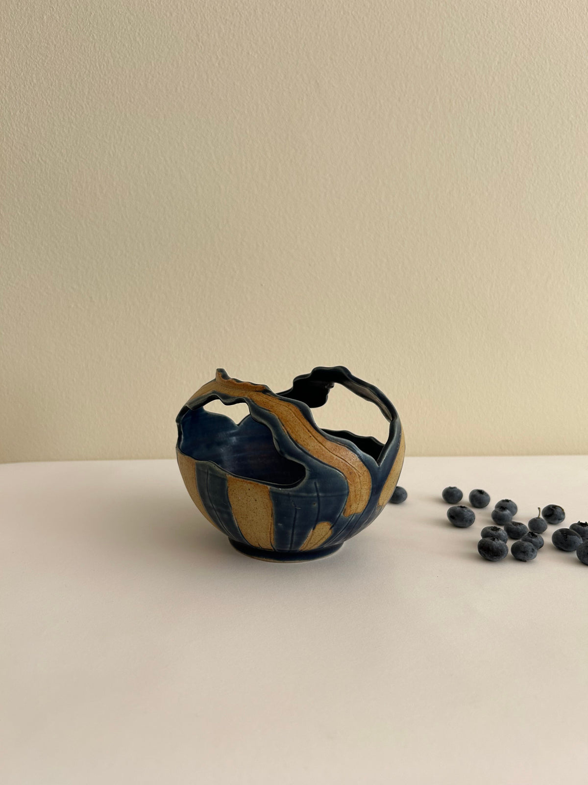 The Artisan Ceramic Bowl by Flock Studio, a small handmade piece with a basket-like shape, showcases blue and brown swirls as it rests on a white surface. Scattered blueberries nearby add a natural touch against the plain cream-colored background.