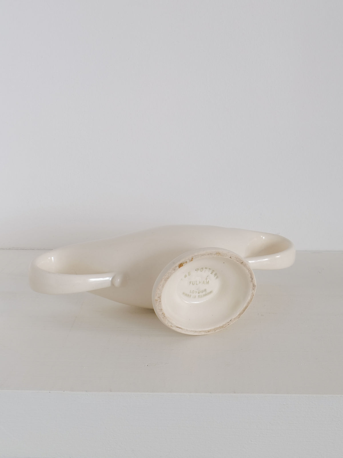 A vintage white ceramic Mini Mantle Vase from the 1930s, by Collection apart, with two handles is lying on its side on a white surface. The vase's base is visible, showing some text imprinted on it. This piece stands out against the plain white background, creating a minimalistic and clean appearance.