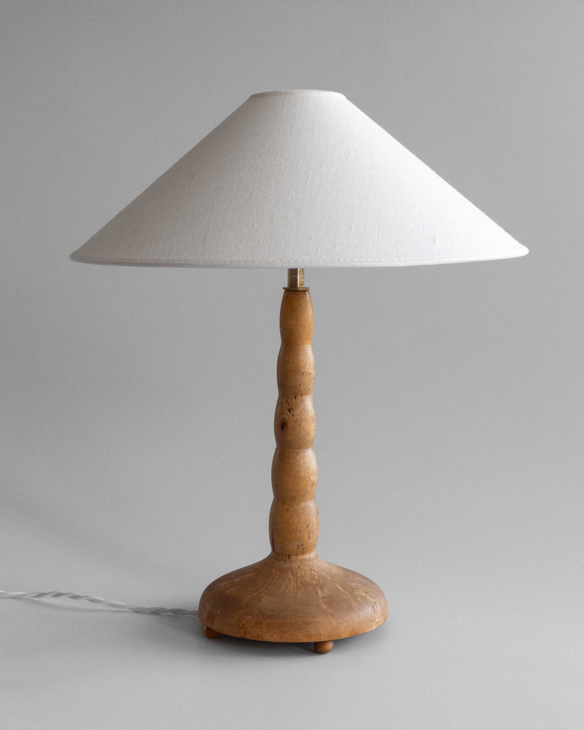 The Spigel Turned Birch Table Lamp from Sweden features a textured wooden rounded base, smooth birch stem, brass fixture, and large white conical lampshade, resting on a neutral background with a visible power cord.