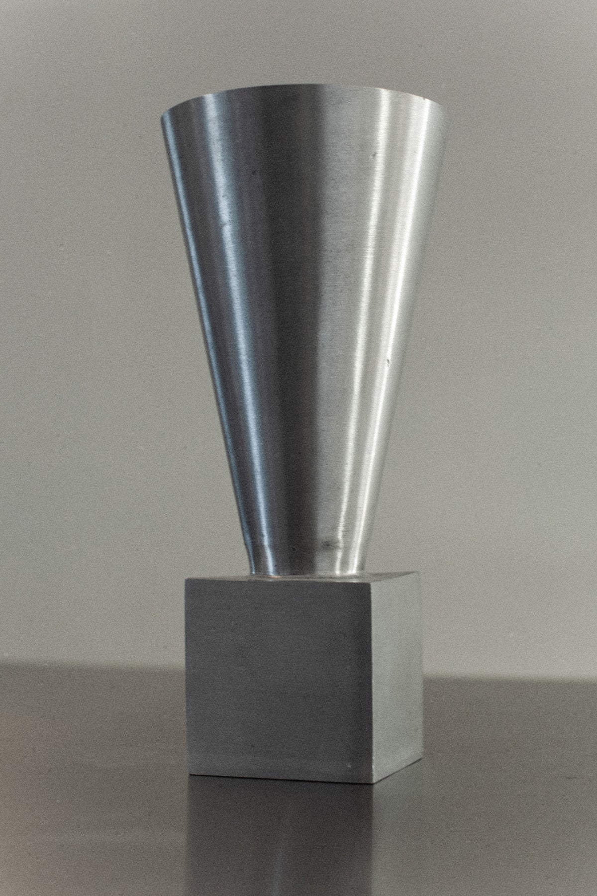 The 70's Aluminum Cast Vase from Out For Lunch rests on a reflective surface against a light gray background. Crafted from brushed aluminum, its conical top and square base blend modern elegance with the allure of minimalist design.