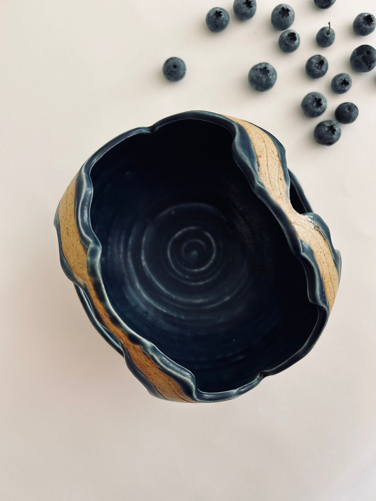 An Artisan Ceramic Bowl from Flock Studio, featuring a wavy rim and basket-like shape, beautifully adorns a white surface. Its dark, textured interior is highlighted by scattered blueberries in the upper right corner, capturing the essence of Balinese artistry.