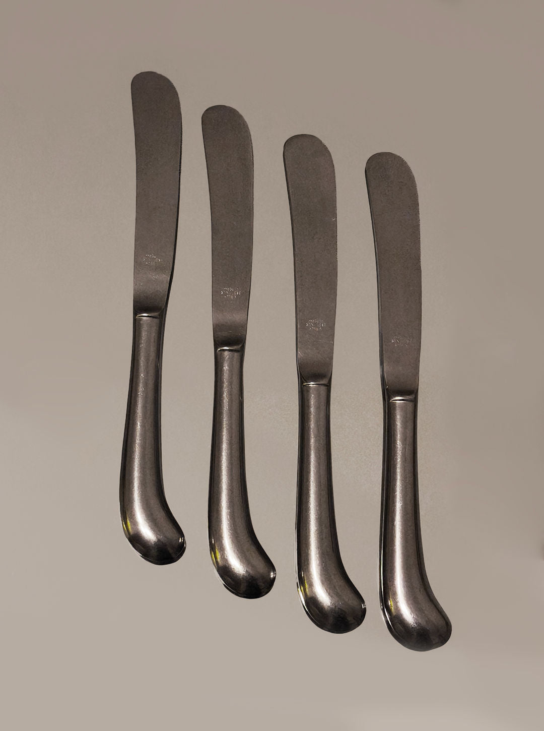 Four Les Objoies stainless steel butter knives from a set of four are arranged vertically side by side on a plain background.