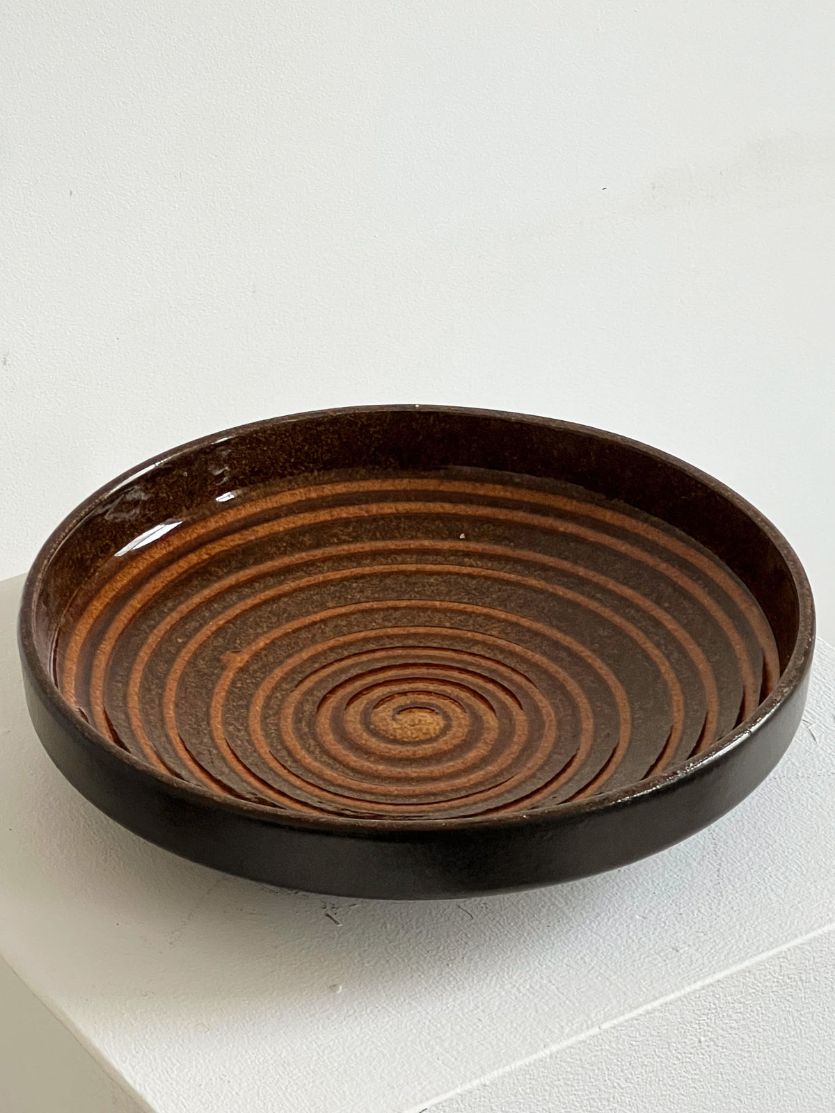 The Porter Studio Spiral Ceramic Dish, a round, dark brown ceramic piece with concentric circular patterns, stands out beautifully on a white background—ideal for kitchen use.