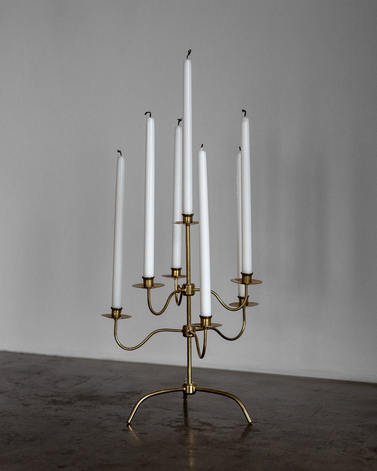 On a gray surface, Spigel's Swedish Mid-Century Brass Candelabra holds six tall white candles, exuding Scandinavian Modern elegance. The blurred background highlights this minimalist piece akin to a Josef Frank design.