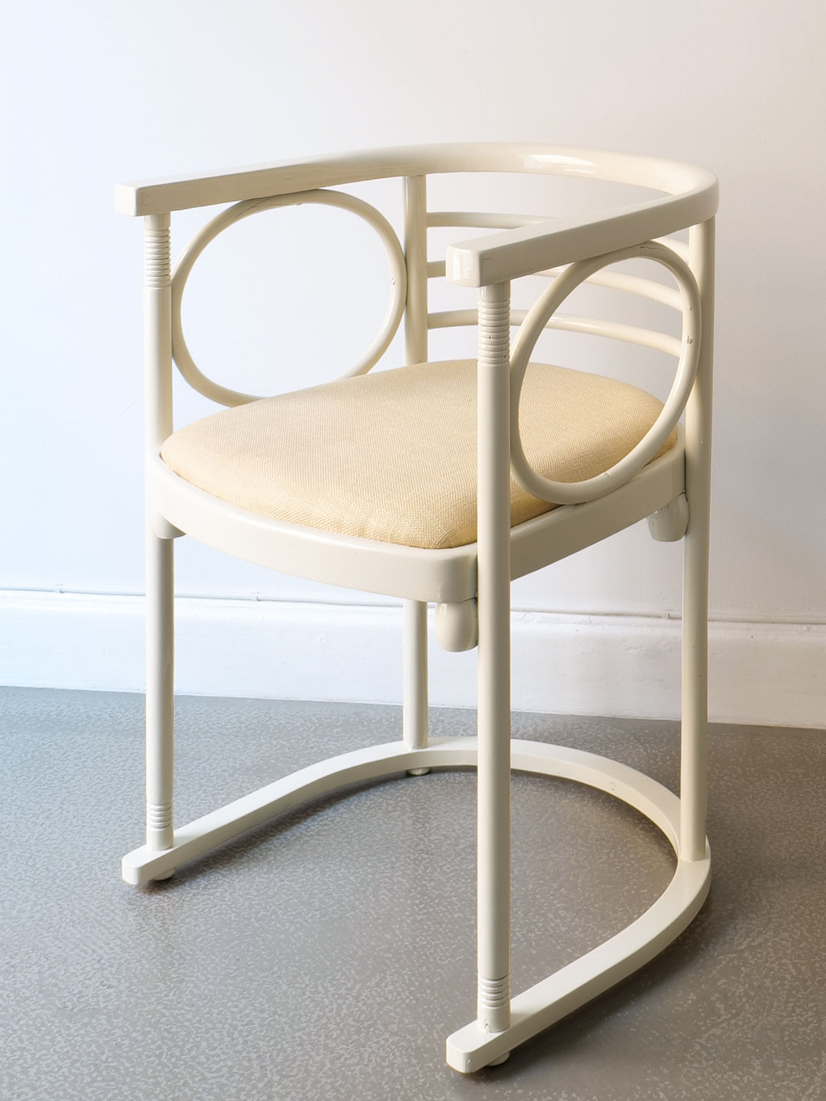 This contemporary chair, from the Collection apart brand's Thonet Bentwood Chairs by Josef Hoffman, features curved lacquered bentwood armrests and a circular back design in a modern cream hue. Its cushioned seat complements the grey floor and white wall, evoking Josef Hoffman's timeless style.