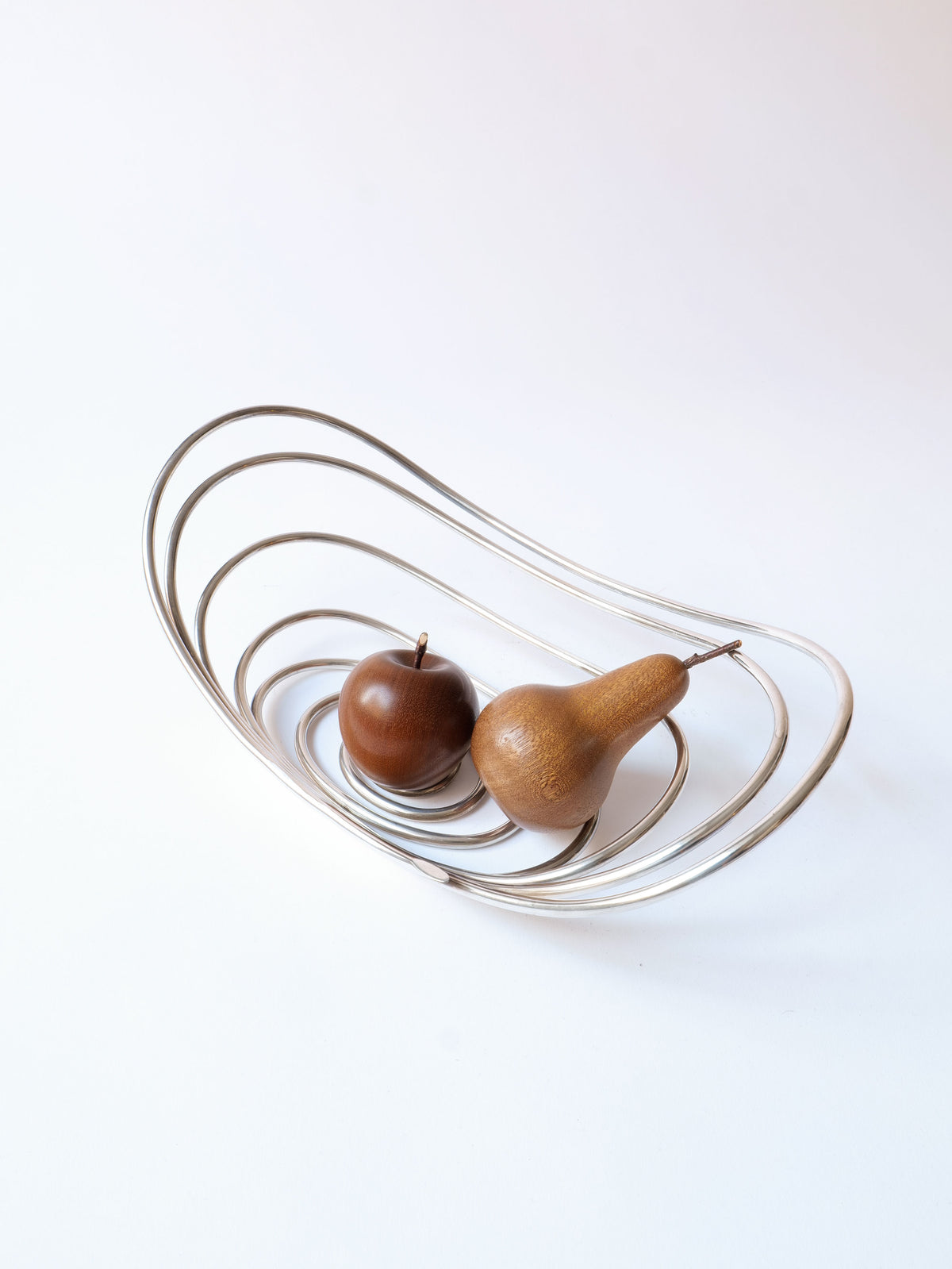 A modernist centrepiece, this minimalist silver wire Centrepiece Lino Sabattini 1970s by Collection apart contains two wooden fruits: one round, resembling an apple, and the other pear-shaped. Designed by an Italian designer, the bowl features smooth, curving lines creating an open, airy structure against a white background. Highly collectible.