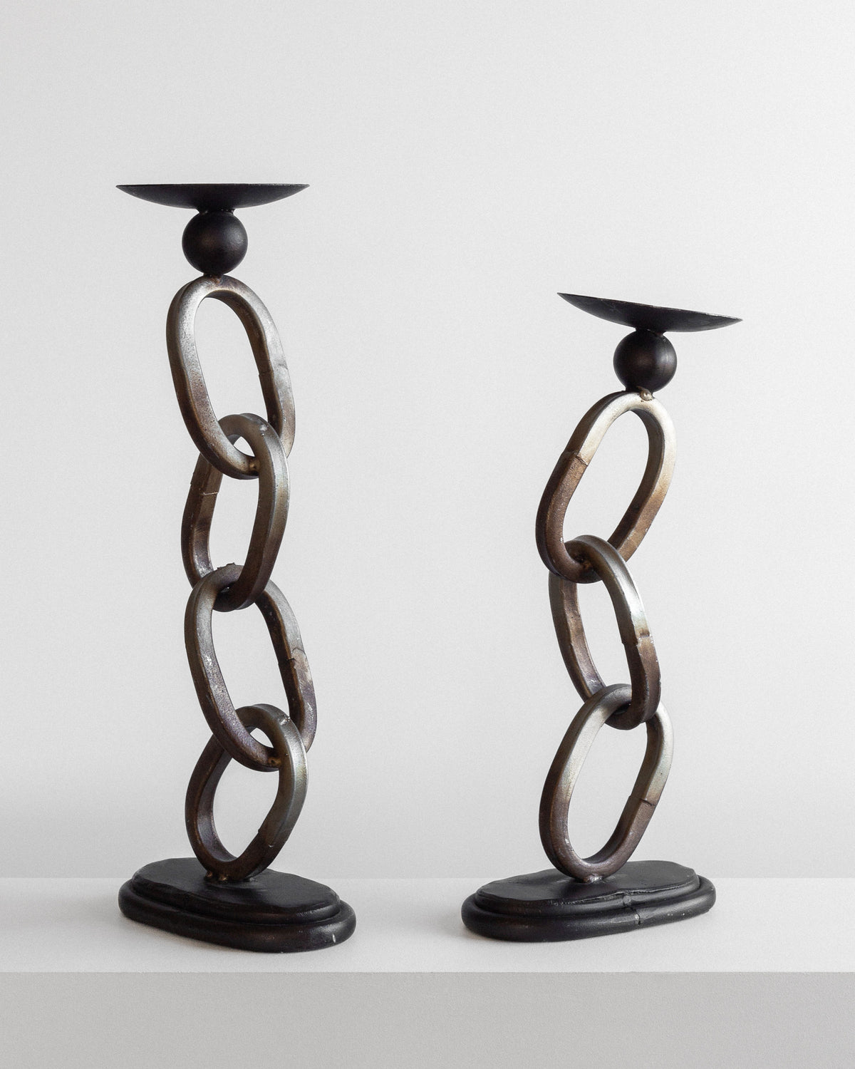 The Postmodern Sculpture Duo by Spigel features two decorative candle holders designed with vertical chains of interconnected metallic rings, adding an industrial touch to your decor. The chains rest on dark, rounded bases and support flat, circular candle plates on top. They are showcased against a plain white background.