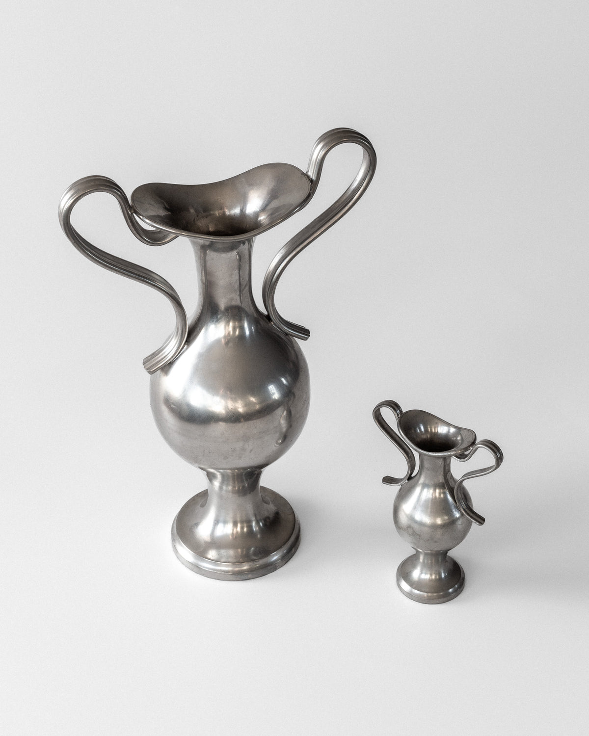 Two pewter neoclassical vases by Spigel, designed by Ernst Svedbom in 1953 Sweden, rest on a flat surface. The larger has a wide curved opening while the smaller mirrors its design. Both have curved handles and a reflective silver finish against a plain white background.