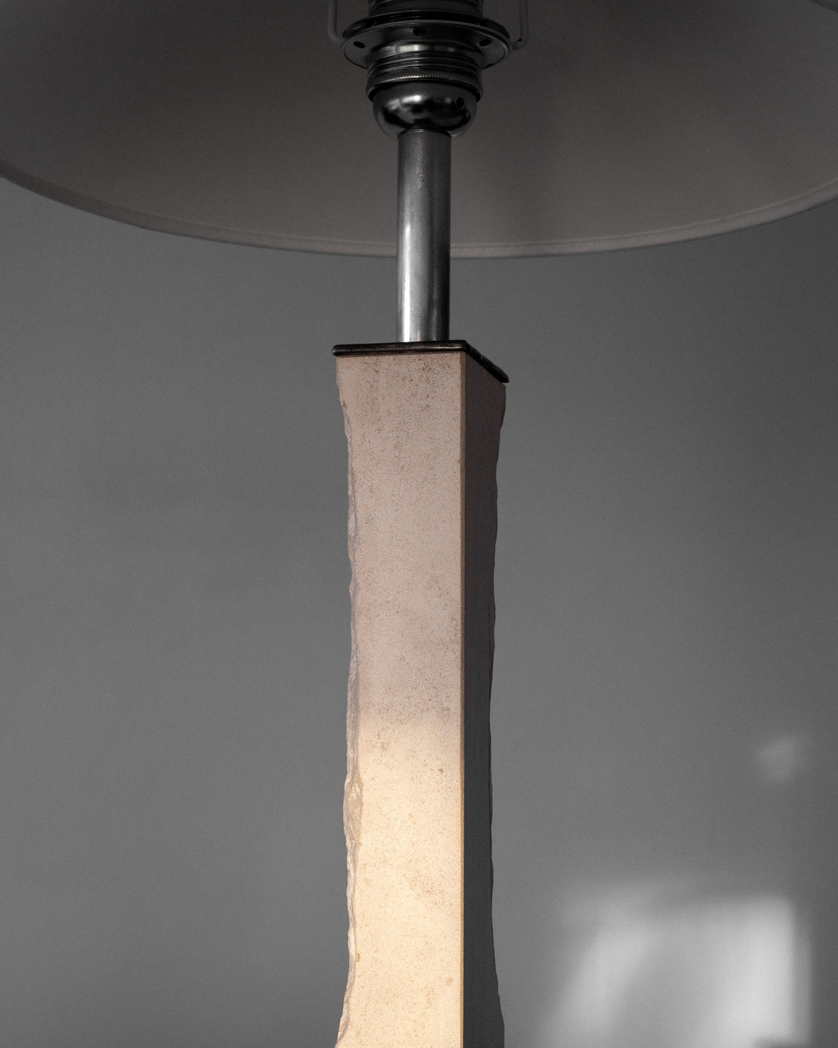 The Spigel Modernist Travertine and Metal Table Lamp showcases a minimalist, mid-century design with a rectangular stone base and metallic neck supporting a simple white shade, casting soft light against a plain gray background.