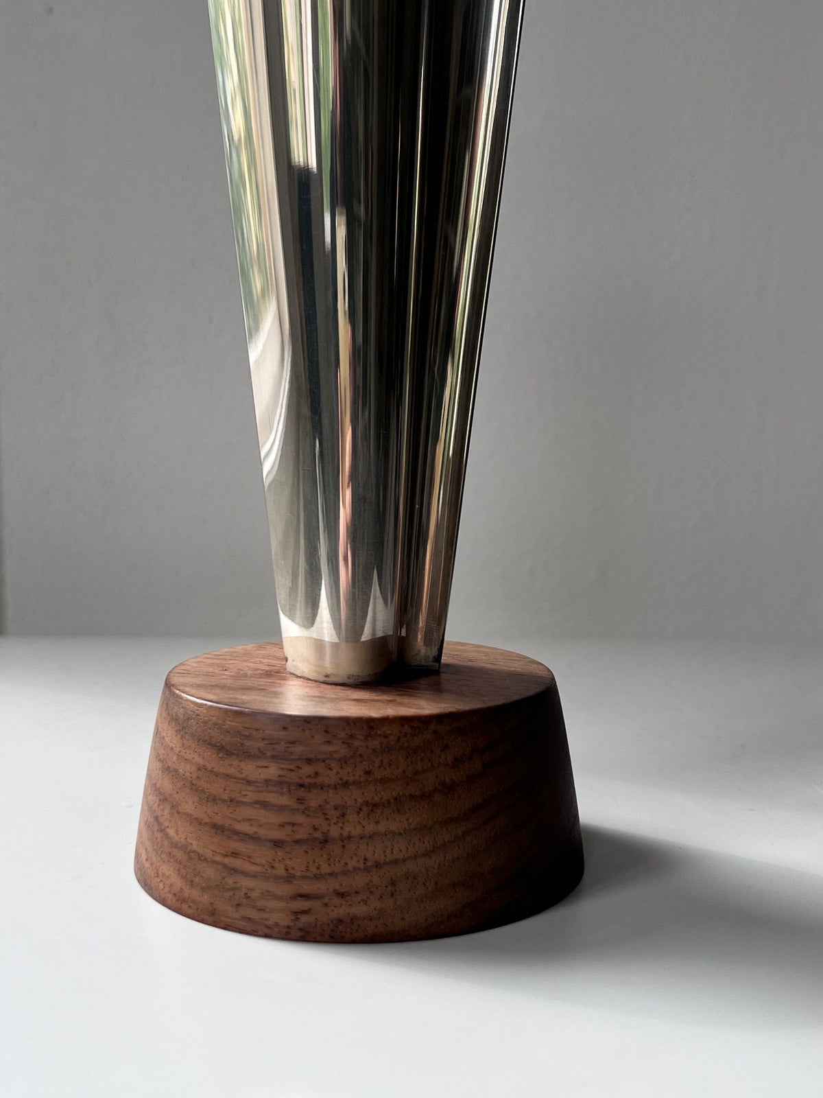 A close-up photo of a polished metal object with a reflective surface, tapered at the lower end, mounted on a circular wooden base. The Médecine Tapio Wirkkala Silver Vase exudes vintage charm against the plain background, with soft lighting highlighting the smooth textures of both the metal and wood.