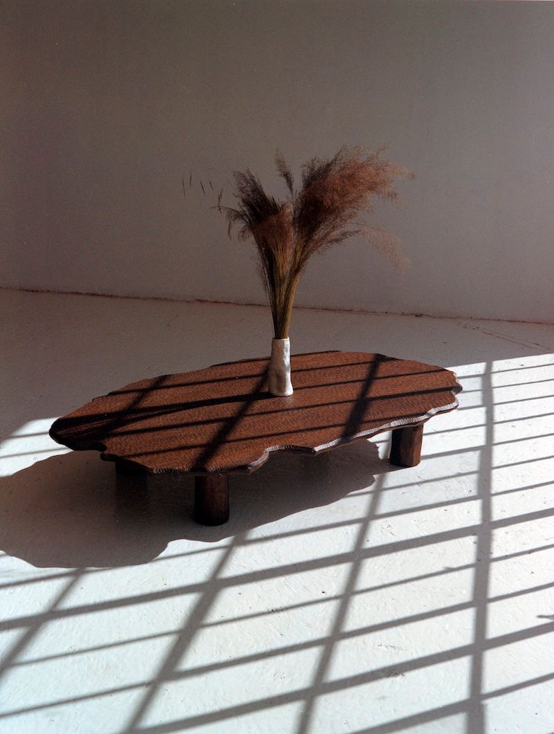 The Mare Coffee Table by Ombia Studio, hand-carved from brown white oak, stands on a white floor with a small vase of dried grasses, sunlight streaming in and casting window bar shadows across the scene.