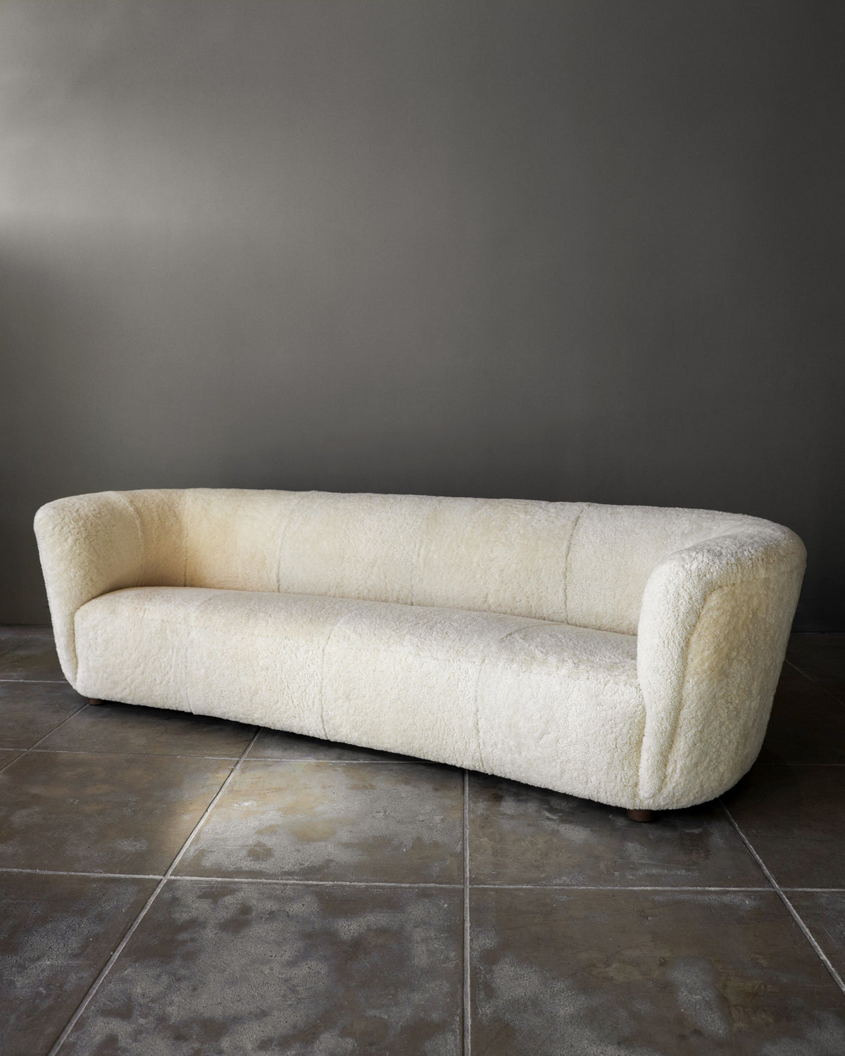 A long, curved Fritz Hansen Sofa in Shearling by Studio OSKLO with a low backrest and rounded arms sits on a tiled floor against a plain gray wall.