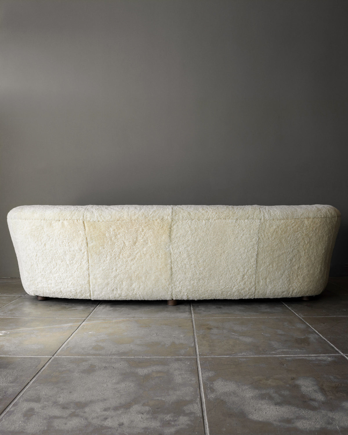 A Fritz Hansen Sofa in Shearling by Studio OSKLO, featuring a long, low-profile cream design with textured fabric, is placed against a plain gray wall on a tiled floor. The image captures the back of the sofa.