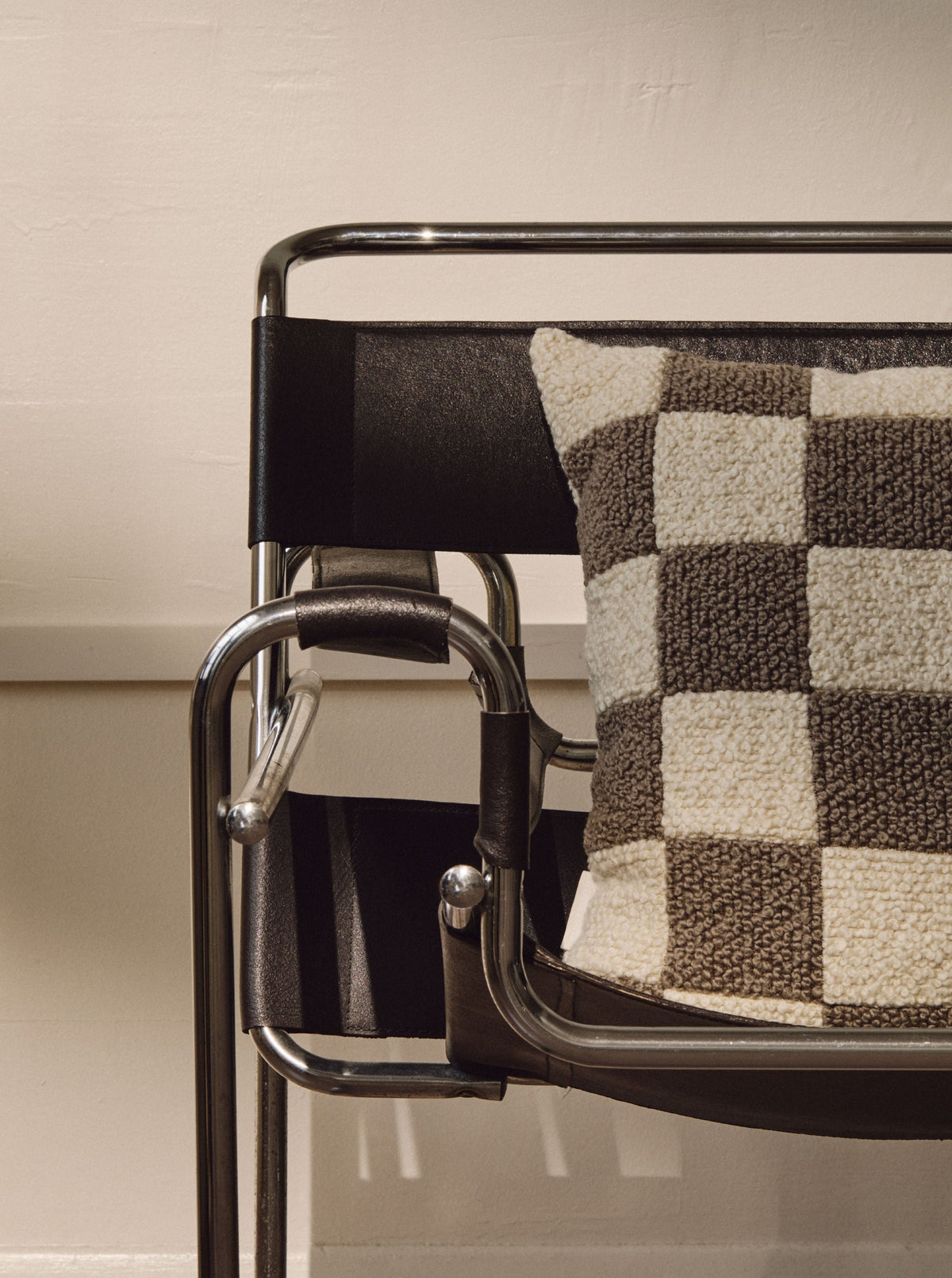 A modern black and chrome chair with a geometric design is displayed against a beige wall. It features AMÉLIORÉ's Le Carré - Wool Bouclé - Sawdust/Eggshell cushion, crafted from British eco wool, adding comfort and vintage-inspired style to the minimalist setting.