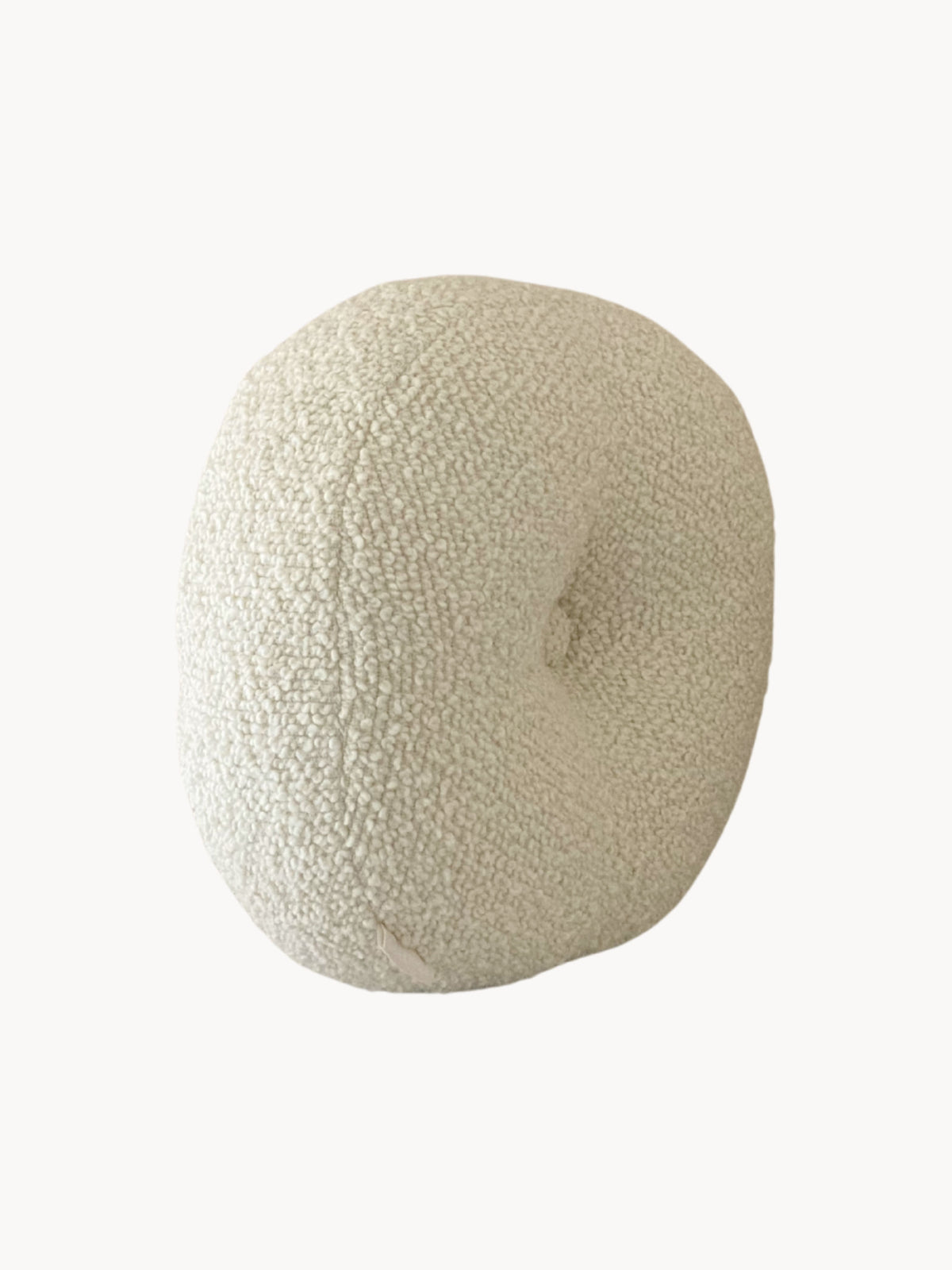 A round, beige, textured pillow crafted from wool bouclé fabric with a dimpled center. The pillow, Le Rond - Moyen - Wool Bouclé - Egg White by AMÉLIORÉ, appears soft and plush, likely providing comfort and support for the back or neck. Its neutral color and unique texture make it a versatile addition to various decor styles.