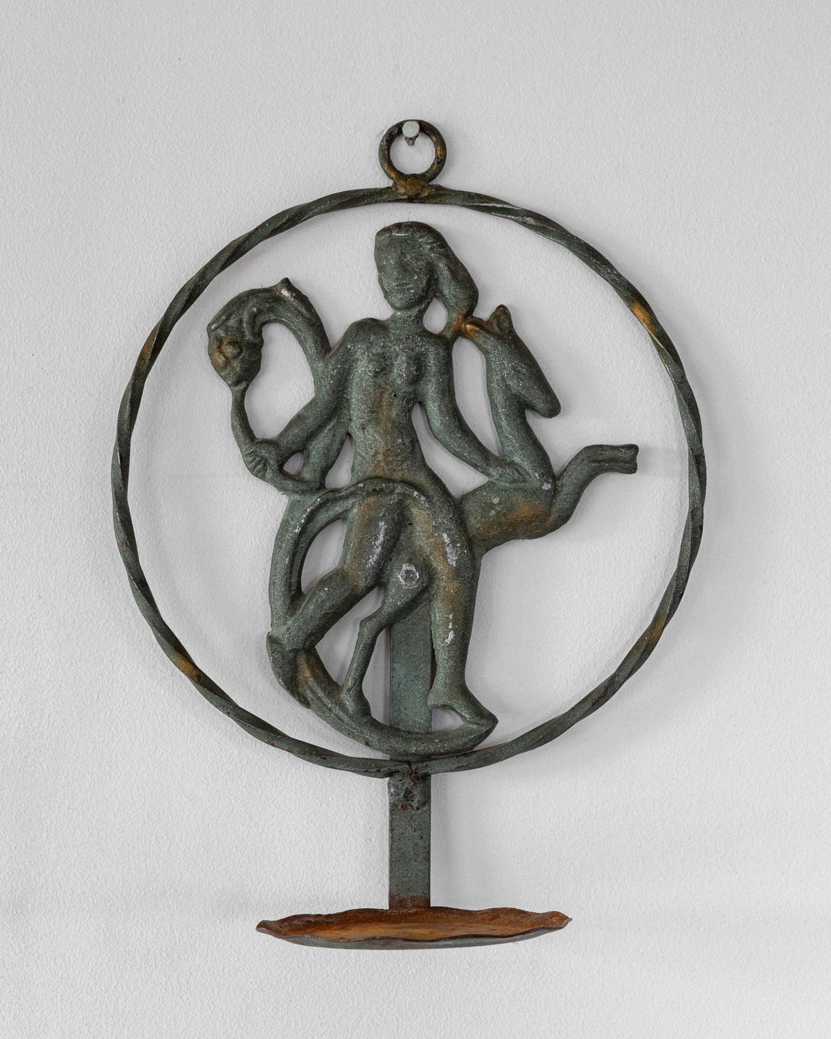The Spigel Art Deco Wrought Iron Wall Sconce from 1920s/30s Sweden elegantly mounts on the wall, depicting a person riding a horse-like creature while holding another figure or animal. The metal's greenish patina adds an antiquated charm to this vintage piece.