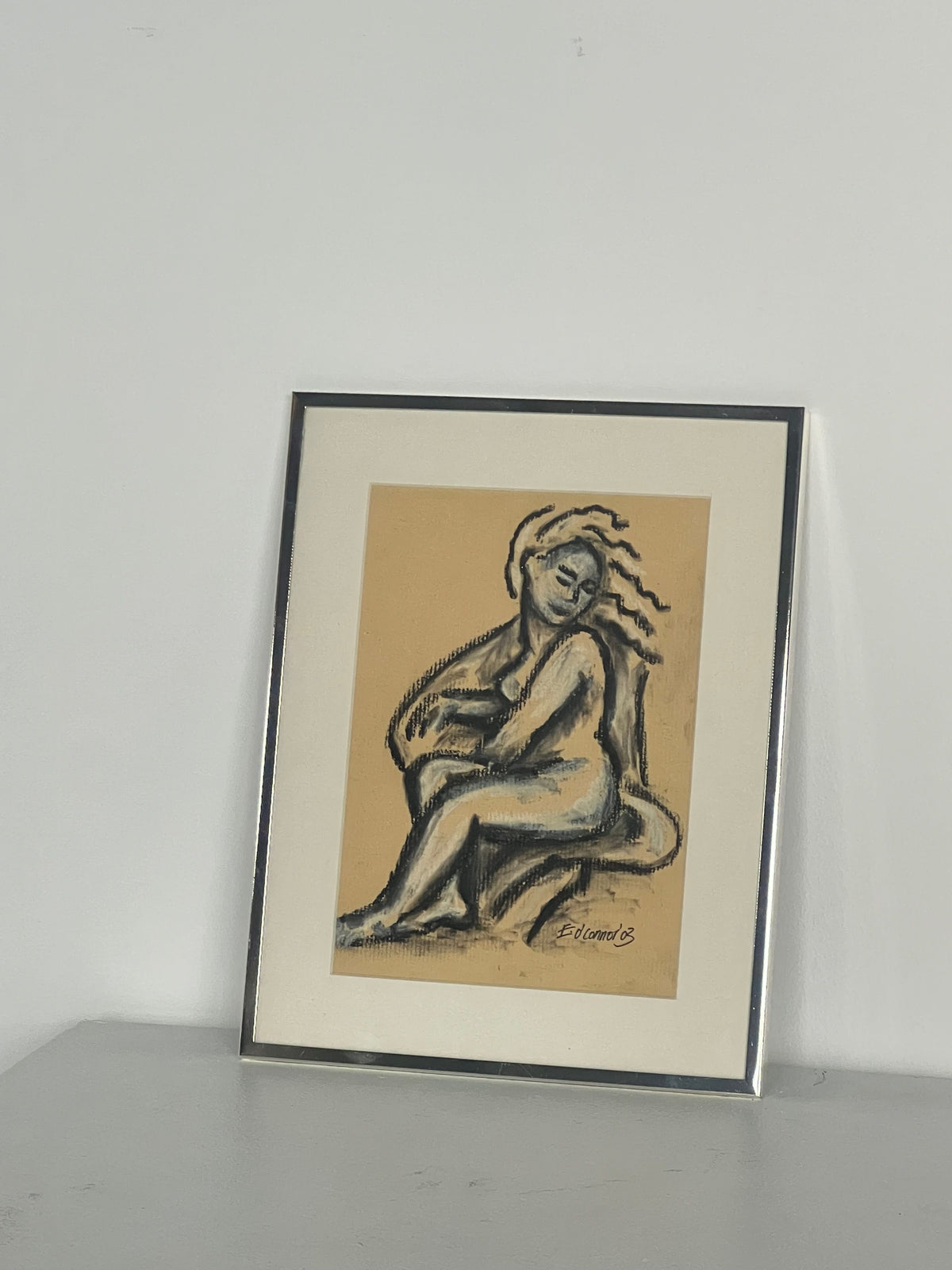 Porter Studio presents "Pastel Drawing" by E.D Connor: a stylized seated figure with flowing hair on beige, set against gray. Framed in thin polished chrome, the artwork is complemented by a white mat.