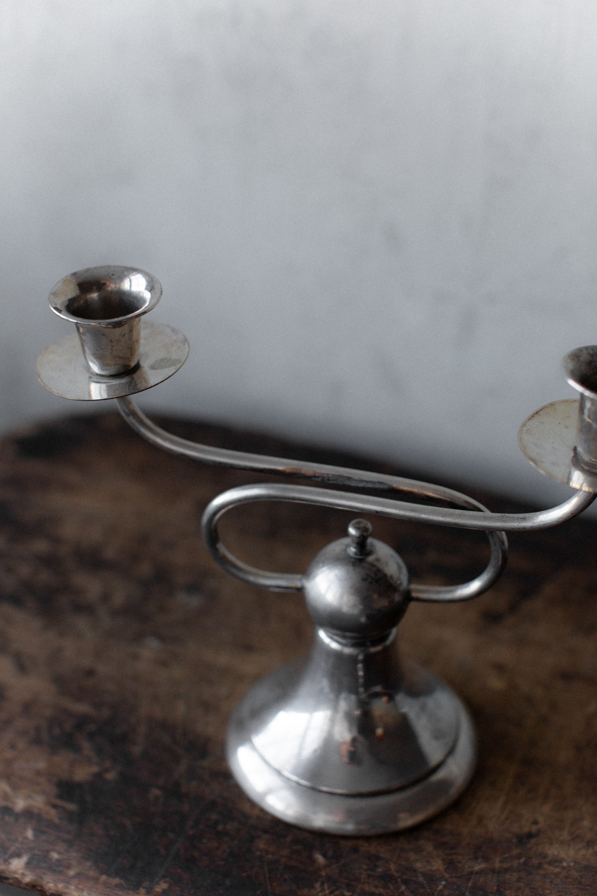 A tarnished silver Art Deco Candleholder by Out For Lunch rests on a rustic wooden surface. With a rounded base, it features two arms extending oppositely, each with a candle socket, embodying geometric elegance against a muted, textured wall.