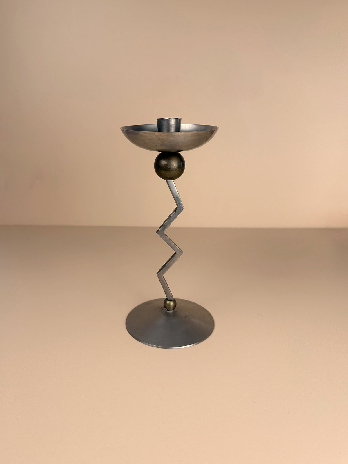 A contemporary, metallic candle holder with an elegant design, the Dodo Vintage Postmodern Zig Zag Candle Holder features a round base, a zigzag-shaped stem, and a shallow circular holder at the top. It is expertly crafted from silver-colored metal against a solid, neutral background.