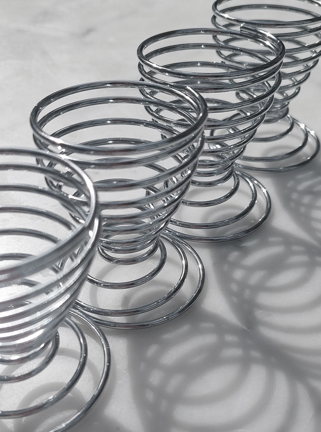 A close-up image showcases a Set of 4 Spirale Egg Cups by Les Objoies with spiral designs, placed in a straight line on a light surface. The egg cup measures create intricate shadows due to the lighting, emphasizing their circular coils and smooth texture.