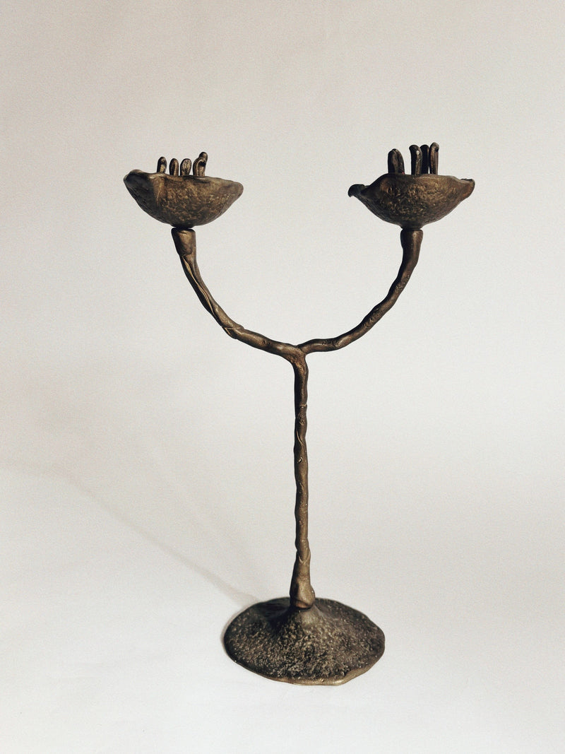 The Huerto 2 Arm Bronze Flower Candelabra, crafted in the 1990s by a Polish artist, features two curved arms ending in hand-shaped cups with upright fingers, all supported by a textured base and set against a plain background.