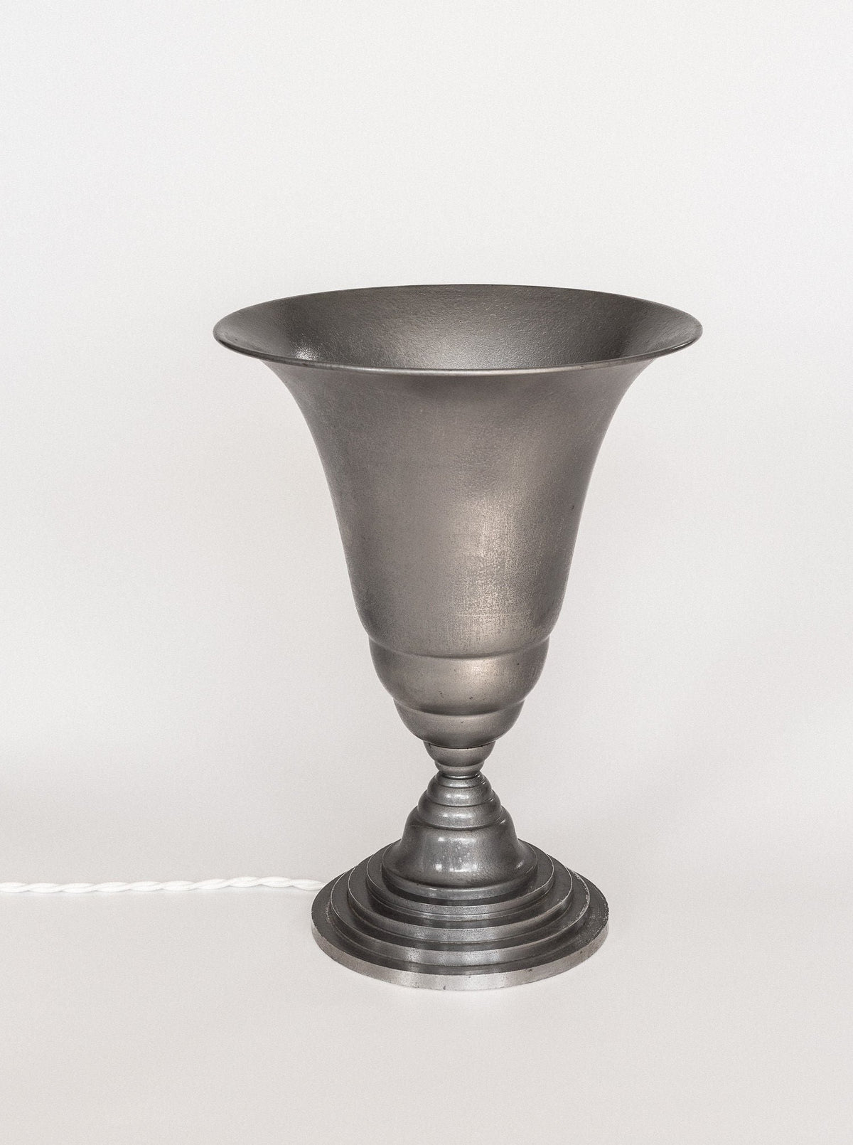 The Art Deco Torchière Table Lamp by Spigel, a gray, flared metal lamp with a round, tiered base, is displayed against a plain white background. The oxidized aluminium lamp showcases a minimalist, industrial design, with a white, twisted cord visible on the left side of the image.