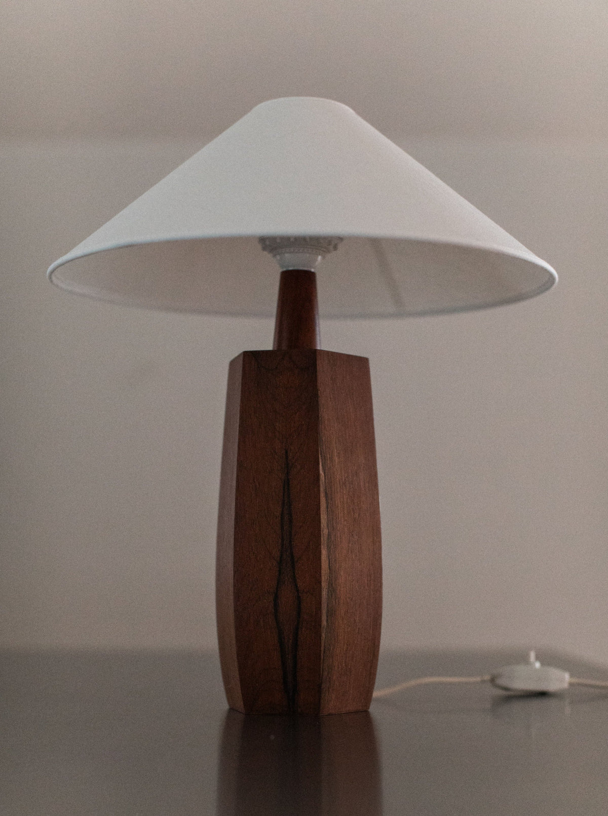 The 1940's Wooden lamp by Out For Lunch exemplifies mid-century design with its wooden, hexagonal base, complemented by a white conical lampshade. Positioned on a reflective dark surface, its meticulous craftsmanship is evident. A white power cord is visible at the base against a plain, light-colored background.