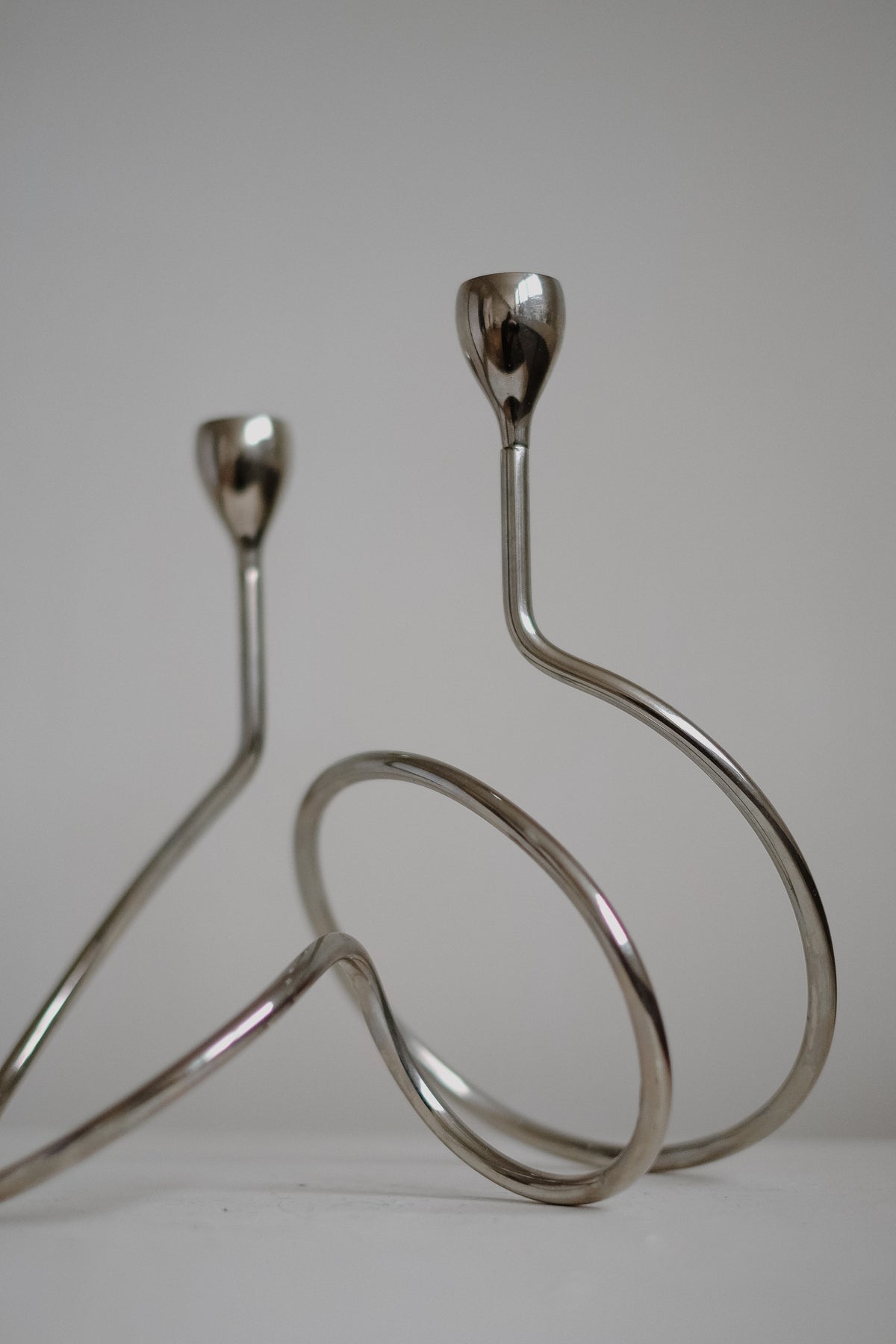 Two sleek "Loop" Candle Holders by septembre studios, with modern chrome sculptural design and looping bases, sit against a neutral background. Their smooth curves lead to simple upward-facing cups ideal for dinner candles.