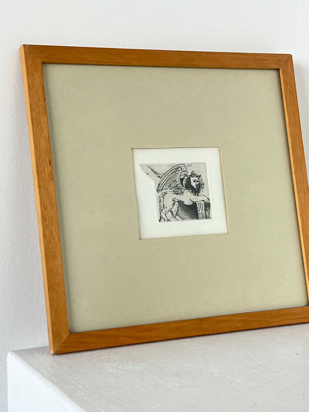 The "Framed And Mounted Hand Illustration" by Porter Studio showcases a detailed depiction of a mythological creature with wings and the body of a lion, enclosed in a small square surrounded by a broader mat border. This intricate illustration in Latin style is encased in an oak frame and elegantly leans against a white wall.