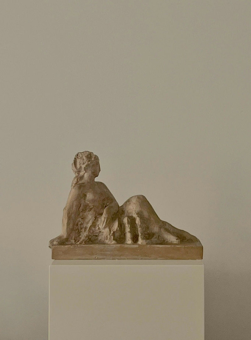 The Sculpture by Sakari Tohka 1938 from Médecine features a small bronze reclining female figure, relaxed and leaning on one arm with legs extended, displayed on a white pedestal against a light beige background.