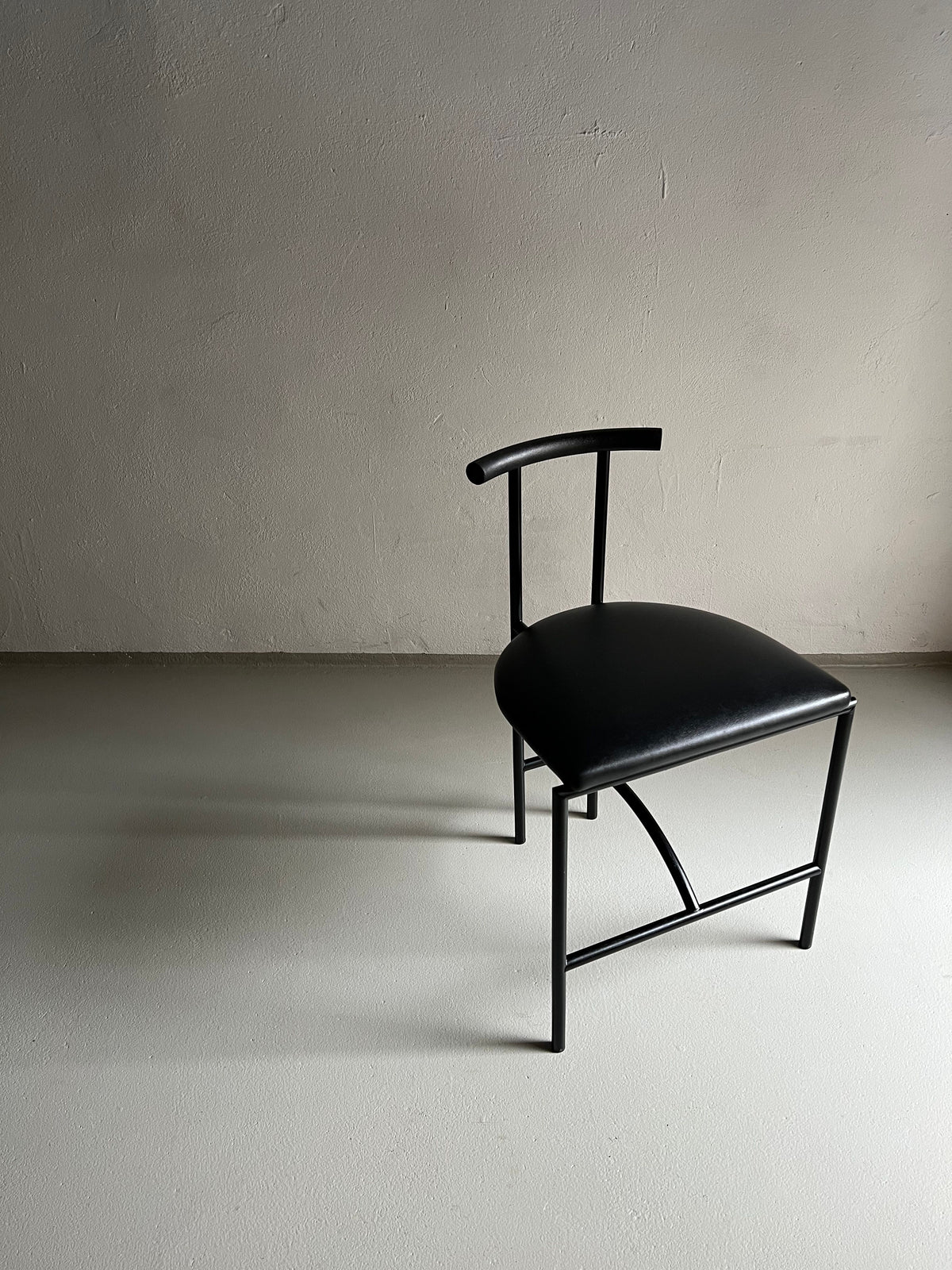 Four Postmodern "Tokyo" Chairs by Rodney Kinsman, produced under the brand Veter Vintage in the 1980s, are placed against a plain, light-colored wall. They have a minimalist black metal frame with thin, curved backrests and faux leather upholstery. The chairs cast soft shadows on the smooth, light grey floor, giving a clean and modern aesthetic to the space.