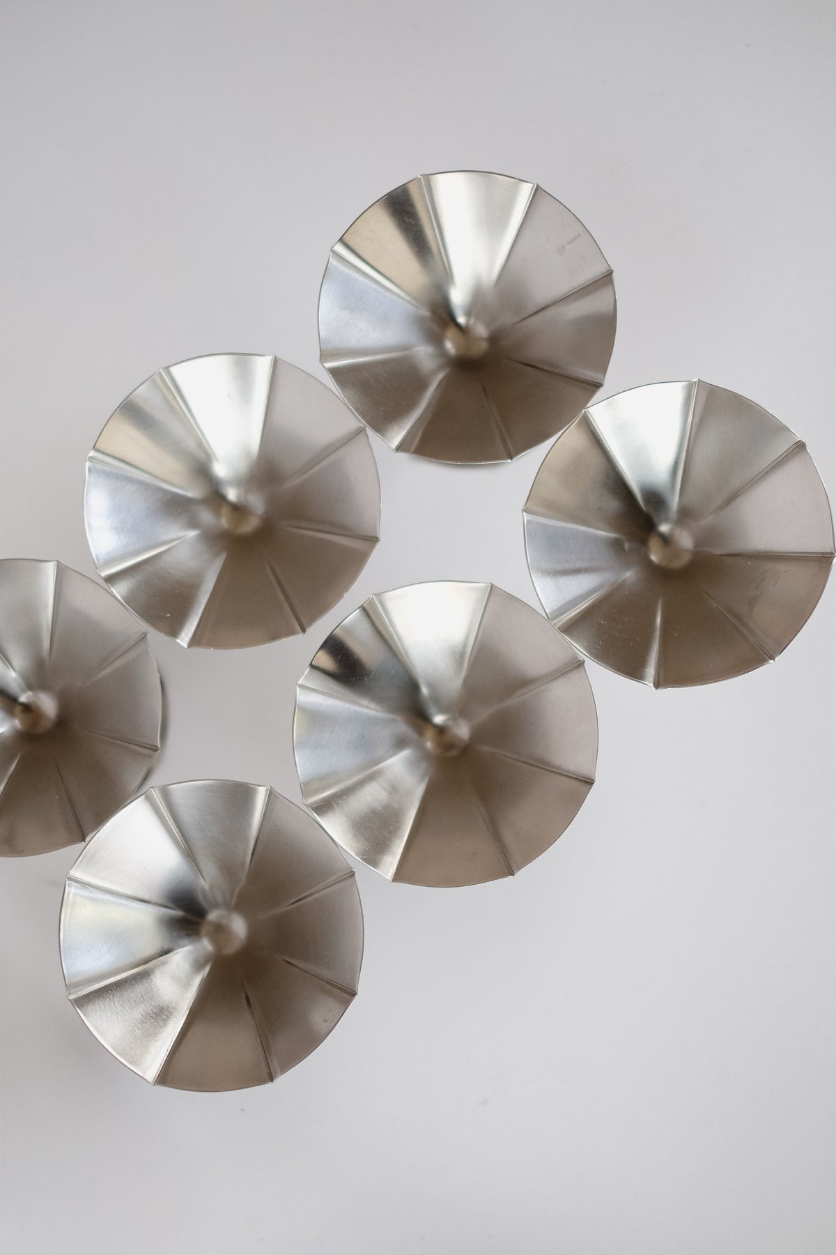 Six metallic, cone-shaped objects from septembre studios' Set of 6 Ice Coupes are arranged in a circle on a light background. Each stainless steel piece has a smooth, shiny surface with a pointed center, creating a visually appealing pattern reminiscent of an ice cream swirl.