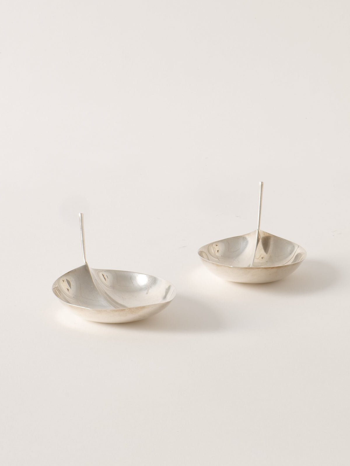 Two modern, minimalist metal serving dishes with sleek, elongated handles in the center. Reminiscent of Tapio Wirkkala Silver Bowls from Collection apart, their smooth, reflective surfaces and clean lines suggest a contemporary style. Placed on a plain white background, they exude the elegance found in prestigious museum designs.