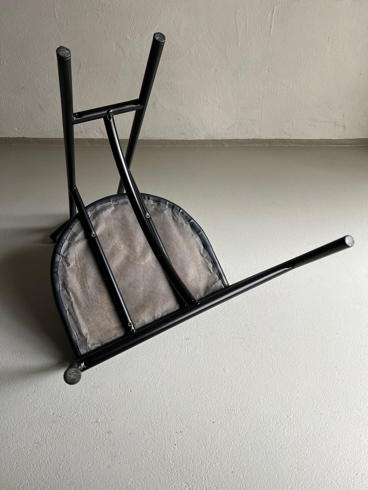 Four Postmodern "Tokyo" Chairs by Rodney Kinsman for Veter Vintage, Italy, 1980s, with black metal frames and faux leather upholstery are overturned on a plain grey floor. The chair legs are visible pointing upward, and the backs of the seats, which appear worn from vintage condition, are resting on the floor. The background is a plain, slightly textured white wall.