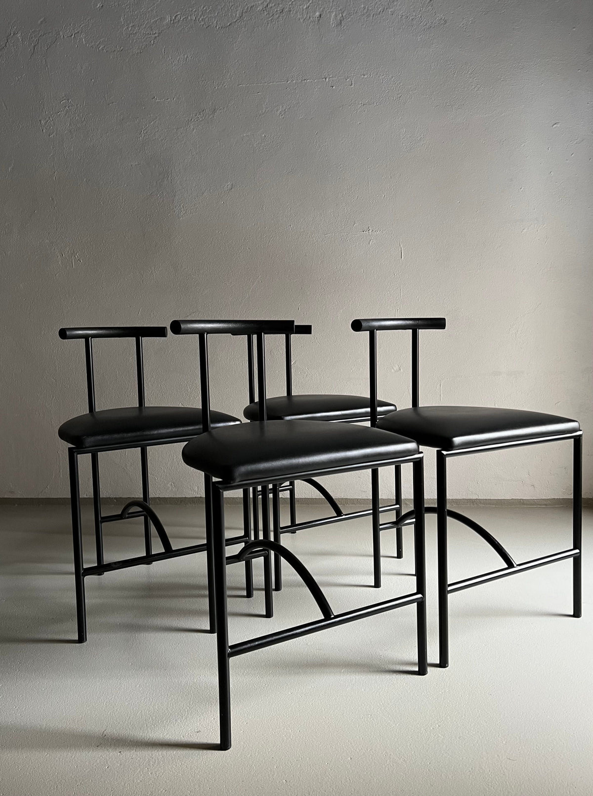 A set of three minimalist black metal chairs with thin frames and T-shaped backrests, featuring faux leather upholstery, is arranged in a staggered formation on a light-colored floor against a plain background. The design is modern and sleek, with a focus on simplicity and functionality. 4 Postmodern "Tokyo" Chairs by Rodney Kinsman | Italy | 1980s for Veter Vintage complete the look perfectly.