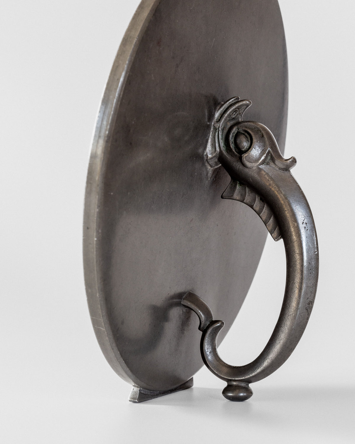 The Spigel Art Deco Metal Mirror, a vintage piece with a round reflective surface, showcases an ornate handle featuring a fish-inspired motif. It stands elegantly against a light gray background, capturing the timeless allure of Art Deco mirrors in perfect vintage condition.