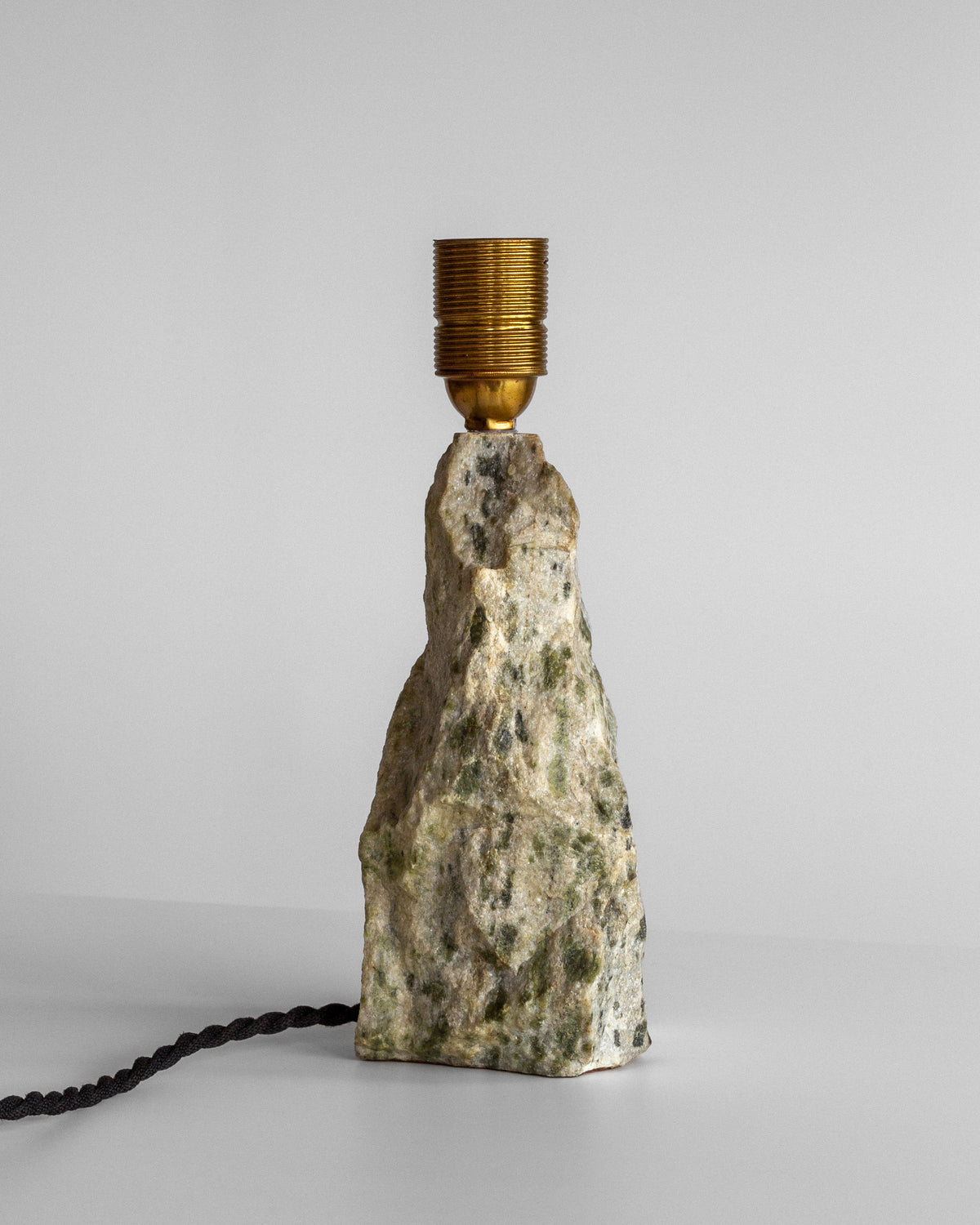 The sleek Marble Stone Table Lamp by Spigel, featuring a minimalist obelisk design with a textured rock base and a metallic gold socket, stands elegantly against a plain gray background. Its sophisticated look is further highlighted by a black woven cord.