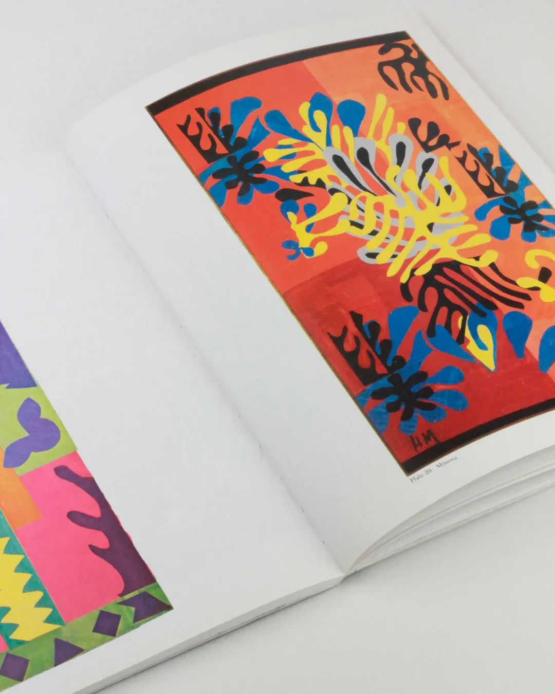 An open book from Boga Avante Shop titled "The Cut Outs of: Henri Matisse" showcases vibrant artwork reminiscent of Henri Matisse's mature cut-outs. The right page features an abstract composition of colorful shapes and patterns in red, blue, and yellow tones, while the left page reveals a corner of another colorful abstract papercutting.